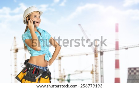 Woman in helmet and tool belt with different tools, talking on phone, smiling. Tower cranes and chimneys in background