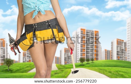 Woman in tool belt holding hammer and pliers. Cropped image. Green hills with road and buildings on background