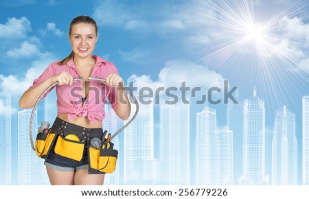 Woman in tool belt with different tools connects two flexible hoses, smiling. Wire-frame buildings as backdrop