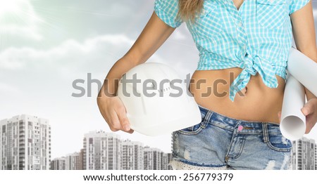 Woman in shirt and short holding helmet and paper in hands. Cropped image. Buildings and sky as backdrop