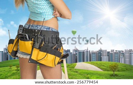 Woman in tool belt with different tools stands back. Cropped image. Green hills with road and buildings on background