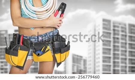 Woman in tool belt with different tools holding cable and pliers. Cropped image. Buildings and gray sky as backdrop