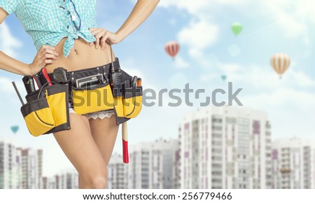 Woman in tool belt with different tools. Hands on hip. Cropped image. Buildings and sky with air balloons on background