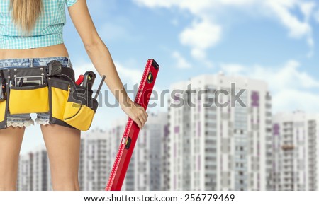 Woman in tool belt with different tools holding red building level. Cropped image. Buildings and sky as backdrop