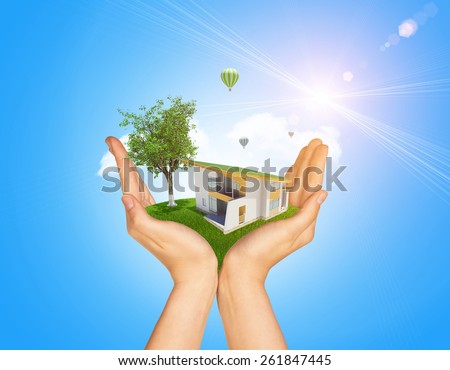 Hands holding cottage on green grass. Background of tree with leaves, hot air balloon and cloud. Blue sky