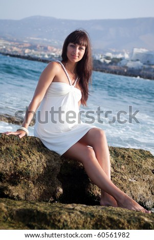 Pretty young female sitting on the rock in front of the ocean shore, looking right in camera. Southern Tenerife, Atlantic Ocean.