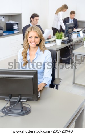 Attractive smiling woman at her desk with handset with people working at the background