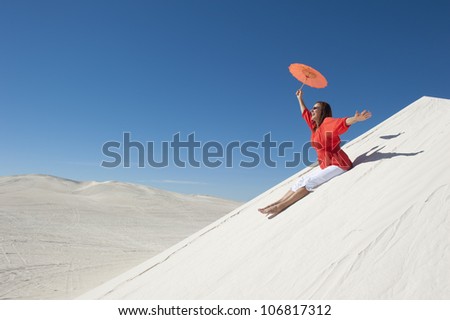 An attractive looking mature woman, wearing red blouse, sitting cheerfully on white sand dune, overlooking a desert scenery and a clear summer blue sky as background and copy space.