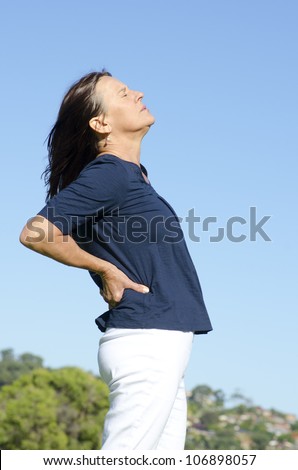 An attractive looking middle aged woman having back problems, feeling the pain in her body, with her eyes closed. Blue sky as background and copy space.