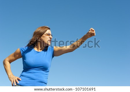 Portrait of angry mature woman, holding up one arm with fist, point towards right side, isolated with blue sky as background and copy space.