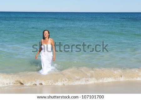 Pretty looking middle aged woman in wet  and soaked white summer dress is coming out of the ocean, isolated with sea and blue sky as background and copy space.
