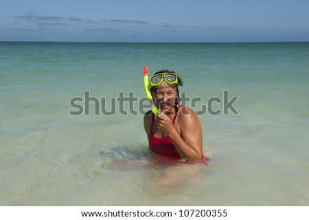 Beautiful looking mature woman in red bathers and yellow snorkel gear, swimming in shallow water of tropical beach, isolated with ocean and blue sky as background and copy space.