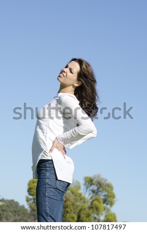 An attractive looking young woman is obviously suffering from back pain, with a stressful facial expression, clear blue sky as background and copy space.