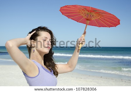 An attractive looking young woman posing relaxed and happy at the beach, holding a red umbrella, with wide open ocean and blurred blue sky as background and copy space.