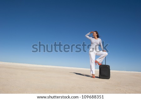 Sexy looking mature woman standing lost in a remote desert area with a suitcase, looking for a transport to travel, isolated with blue sky as background and copy space..