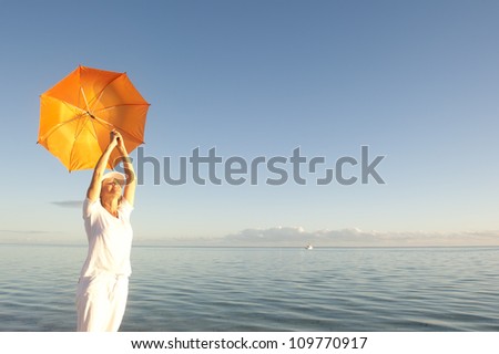 Attractive looking senior woman holding orange umbrella over her head, standing isolated with peaceful ocean and blue sky at sunset as background and copy space.