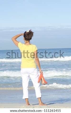 Attractive woman standing alone at beach looking over ocean, with high heel shoes in hand, isolated with sea and sky as background and copy space.