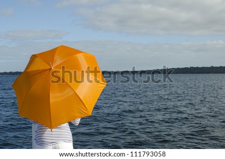 Anonymous woman finds shelter under bright orange umbrella standing at dark water of lake, with coastline and cloudy sky as background and copy space.