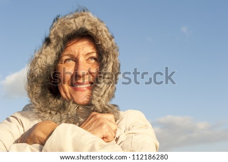 Portrait of attractive looking senior woman keeping herself warm in cold winter weather, isolated with furred hood covering head and blurred background as copy space.