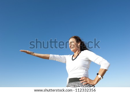 Attractive and confident mature woman holding arm up, proposing, offering. Isolated with blue sky as background and copy space.