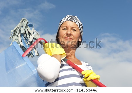 Portrait mature cleaning lady outdoor at spring time, with bucket, mop and bandana, isolated with blue sky as background and copy space.