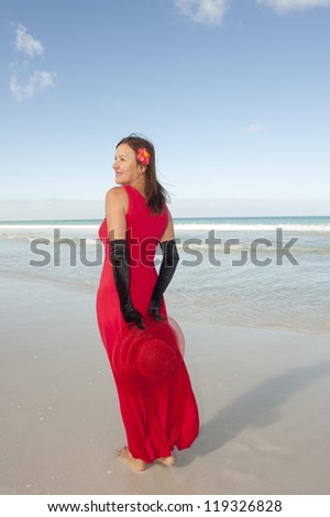 Beautiful looking lady posing happy smiling in elegant red dress and long black gloves at beach, isolated with ocean and blue sky as background and copy space.