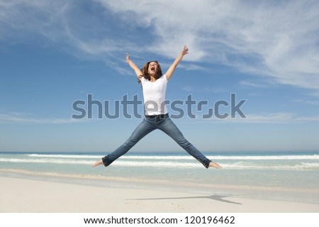 Happy cheerful young pretty woman jumping full of joy at beach with arms up, isolated with ocean and blue sky as background and copy space.