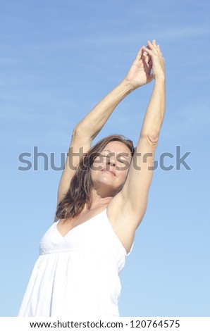 Portrait attractive mature woman outdoor in white summer dress and arms up, relaxed with closed eyes and happy smile, isolated with blue sky as background.