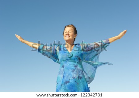 Portrait elegant middle aged woman sunny day outdoor, happy smiling, closed eyes, with arms up and isolated with blue sky as background and copy space.