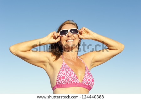 Portrait attractive mature woman in pink bathers and sunglasses posing with arms up happy and joyful at sunny holiday vacation, isolated with blue sky as background and copy space.