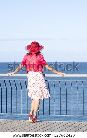 Portrait sexy attractive mature woman dressed up in red, with hat, blouse, dress and high heels, standing relaxed at sunny day at lookout, with ocean and sky as background and copy space.