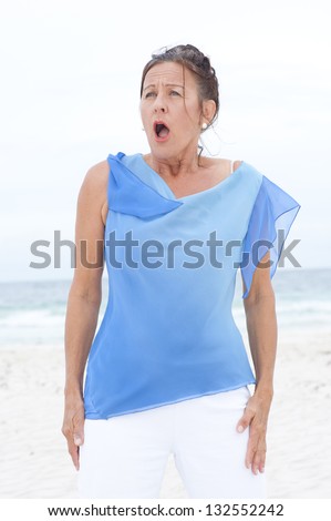 Portrait Beautiful mature woman surprised and upset at beach, wearing blue blouse, with ocean and white overcast sky as blurred background and copy space.