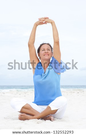 Portrait Beautiful mature woman sitting joyful and happy with arms up at beach, wearing blue blouse, with ocean and white overcast sky as blurred background and copy space.