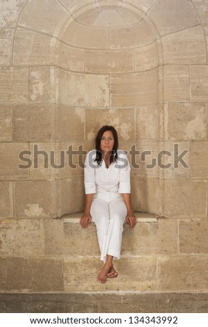 Portrait attractive mature woman sitting alone, lonely and with sad facial expression in alcove at big brick wall.