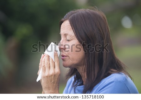 Portrait beautiful mature woman suffering from cold or flu infection, sneezing into tissue, painful seasonal hayfever, with blurred outdoor background and copy space.