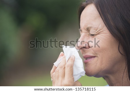 Portrait smiling attractive mature woman suffering from cold or flu infection, sneezing into tissue, painful seasonal hayfever, with blurred outdoor background and copy space.