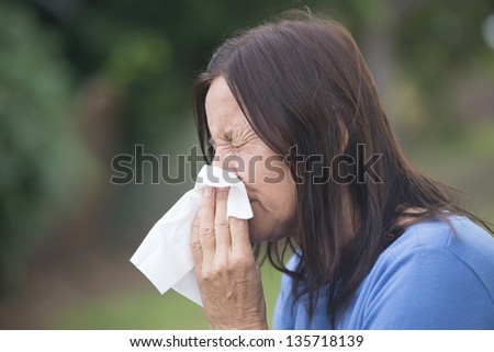 Portrait attractive mature woman suffering from cold or flu infection, sneezing into tissue, painful seasonal hayfever, with blurred outdoor background and copy space.