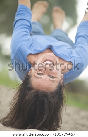 Portrait attractive mature woman hanging bare feet upside down on swing on playground, enjoying cheerful active retirement, happy smiling, with blurred outdoor background.