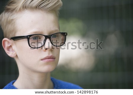 Portrait blond teenage boy wearing glasses with serious face, blurred background.