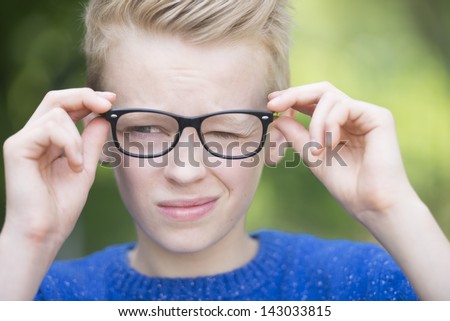 Portrait smart looking blond teenager with glasses and a blink of an eye, thoughtful and clever, outdoor with green blurred background.