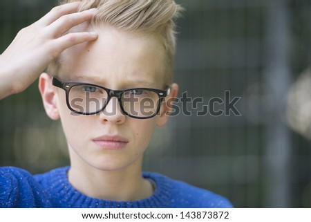 Portrait confident blond teenage boy wearing glasses with serious face, blurred background.
