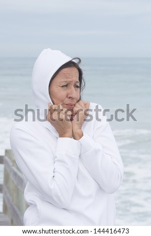 Portrait attractive mature woman wearing white jumper with hood, freezing in cold seasonal autumn or winter weather at ocean, with blurred background of sea.