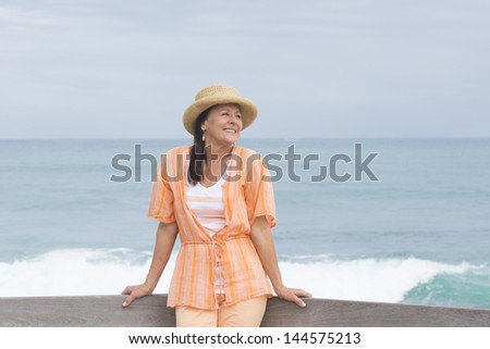 Portrait beautiful mature woman standing relaxed and friendly smiling at beach, wearing orange blouse and hat, with ocean and horizon as blurred background and copy space.