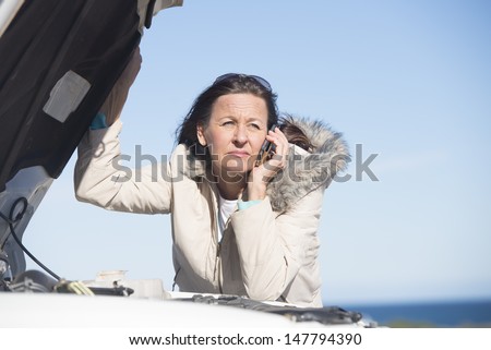 Portrait stressed mature woman breakdown with car calling for service, assistance for help on mobile phone, with blue sky as background and copy space.