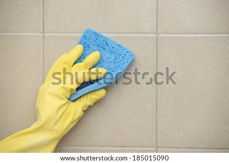 Hand in yellow rubber glove cleaning tiles with blue sponge, copy space.