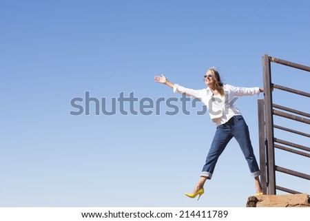 Portrait beautiful mature woman, confident, joyful, happy, with arm up, leg up, wearing high heel shoes outdoor, with blue sky as background and copy space.
