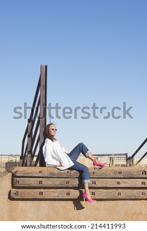 Portrait relaxed attractive mature woman sitting laid back in rural country area, wearing sunglasses, jeans and high heel shoes, with horizon and blue sky as background and copy space.