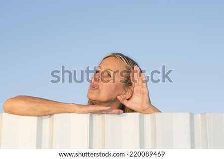 Portrait of attractive mature woman listening curious to noise or sound outdoor, while leaning over metal fence with one hand to ear, with blue sky as background and copy space.