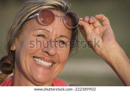 Portrait of happy attractive mature woman outdoor, joyfully smiling, relaxed and laid back facial expression, holding up sunglasses with one hand, blurred background.