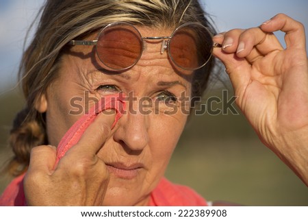 Portrait of attractive mature woman with sad and facial expression, looking concerned, wiping tears or dirt from eyes with tissue, holding glasses up with one hand, with blurred background outdoor.